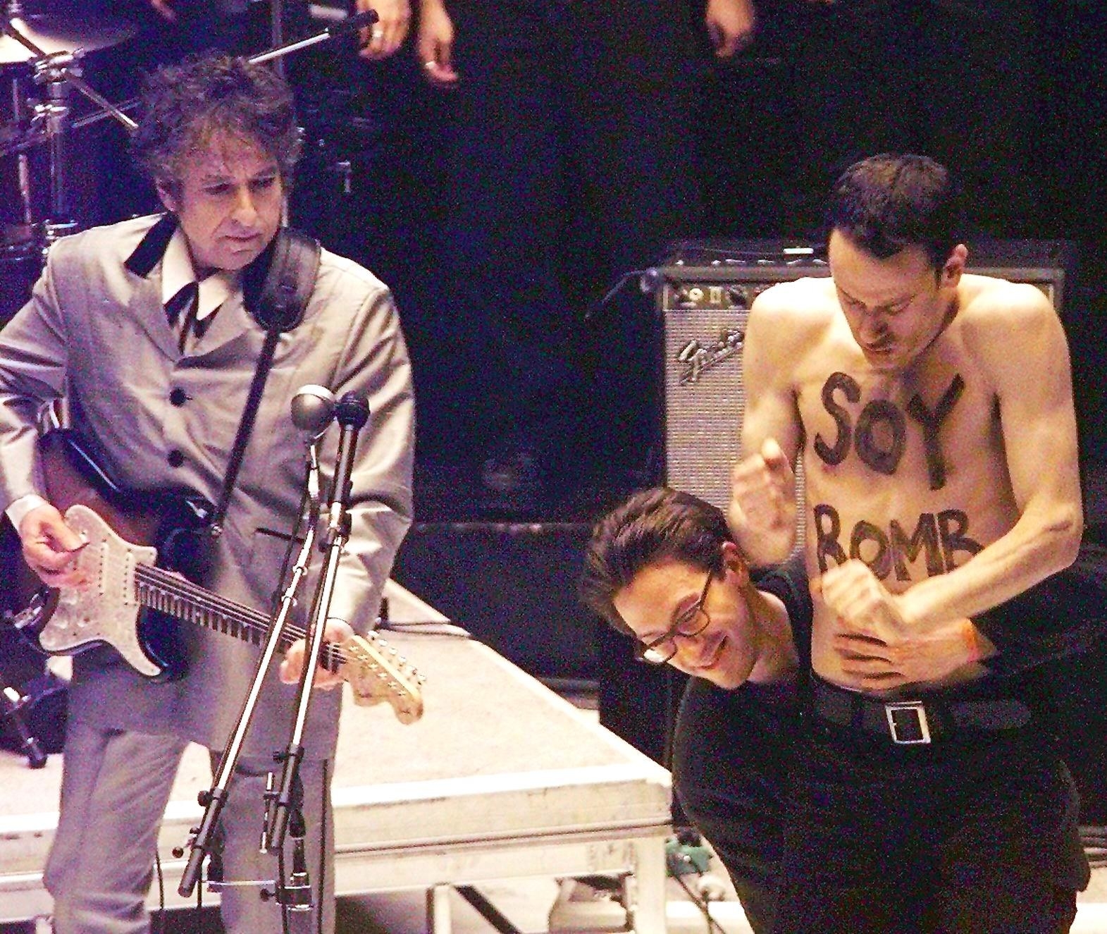 Bob Dylan holds a guitar and watches security take away a shirtless man with Soy Bomb painted on his chest