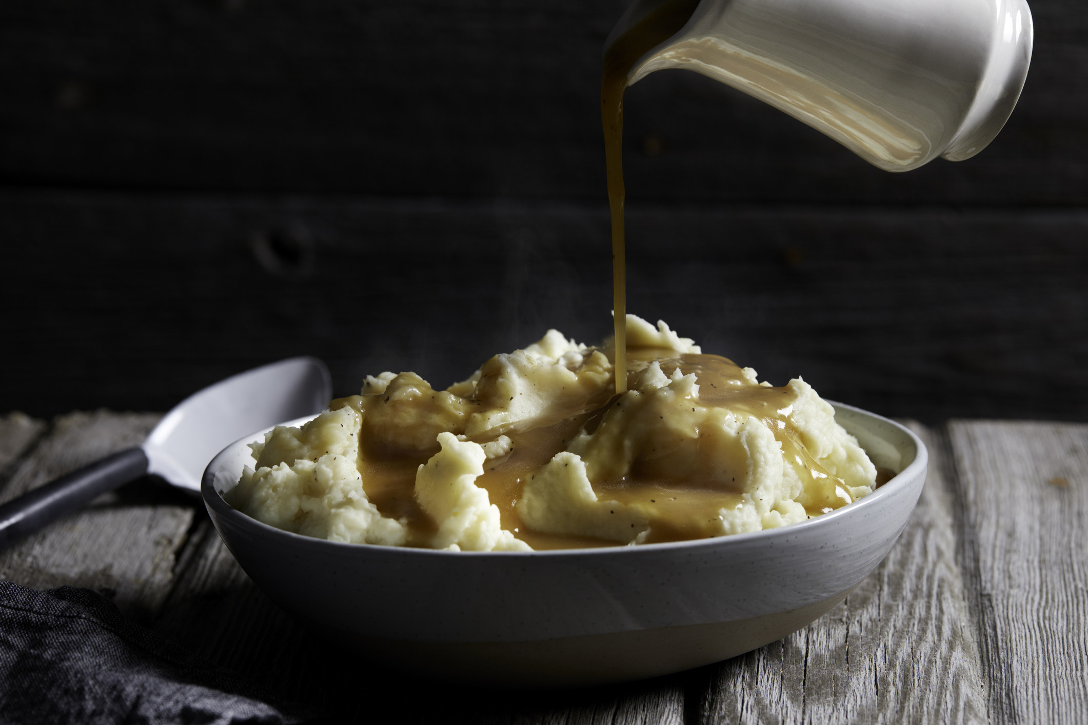 A jug of gravy being poured onto a bowl of steaming mashed potatoes