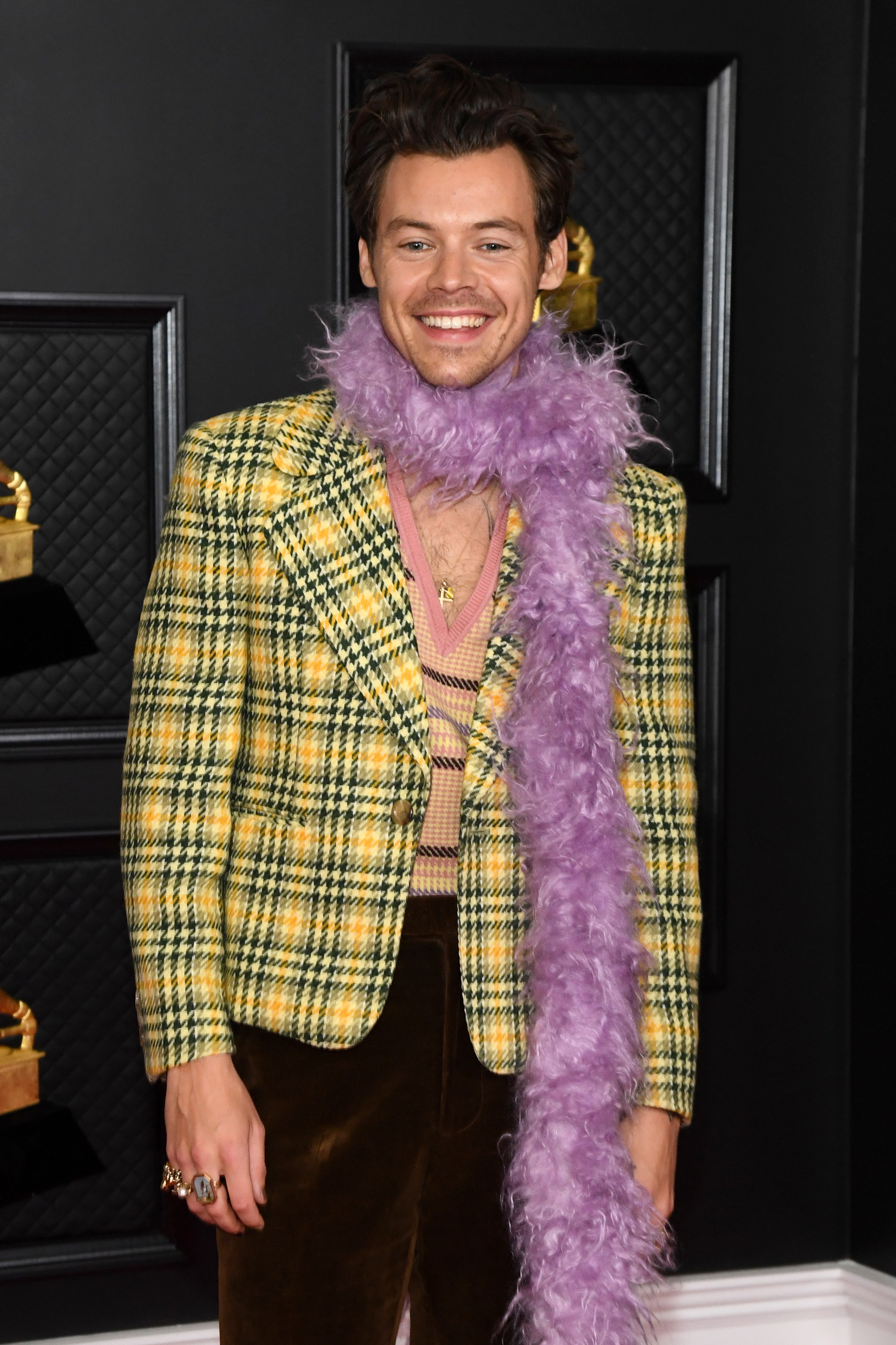 Harry wears a yellow plaid jacket over a knitted V-neck patterned vest and brown cord trousers; he tops it off with a gold necklace and a lilac feather boa