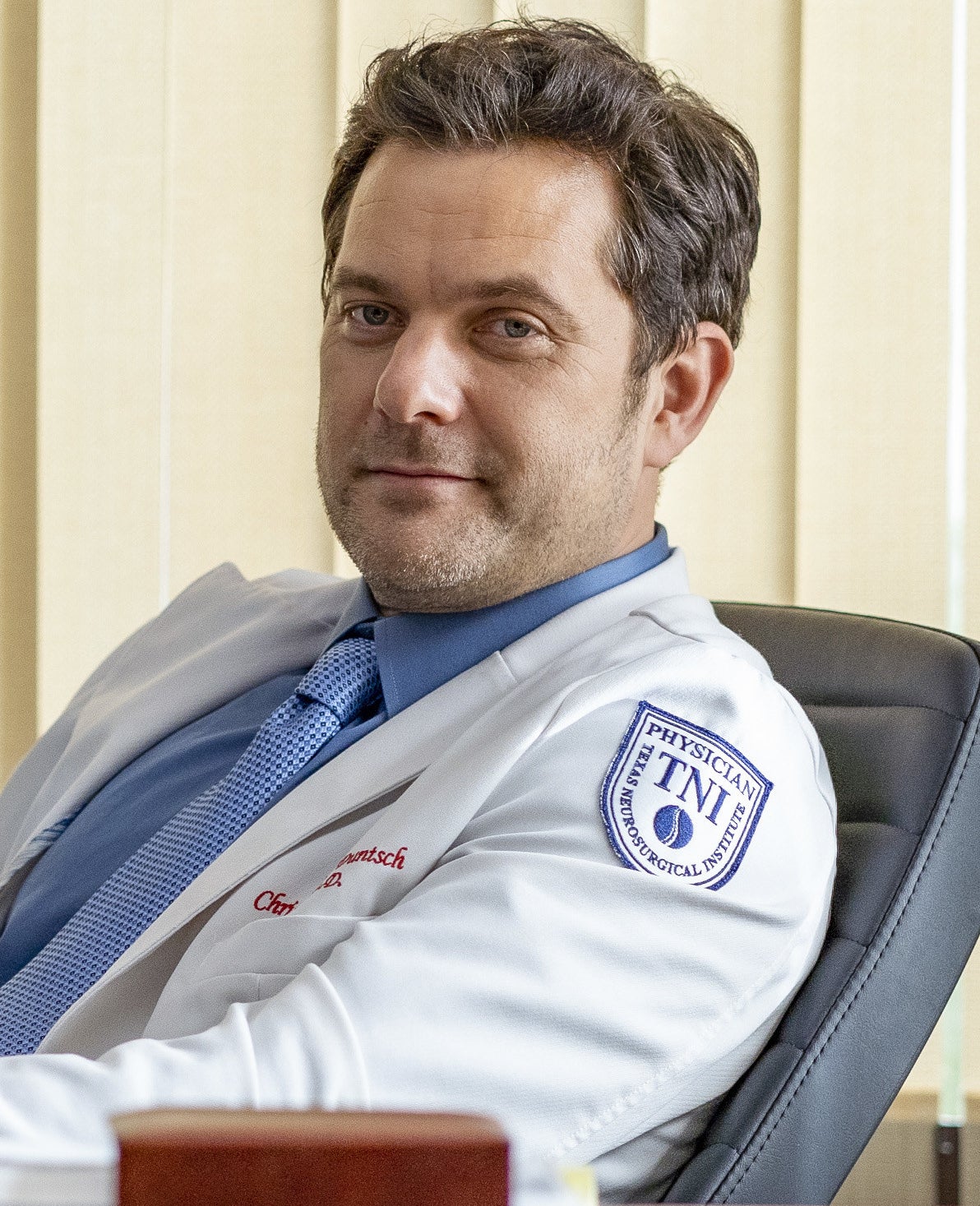 Joshua in an office chair with short hair wearing a physician&#x27;s coat
