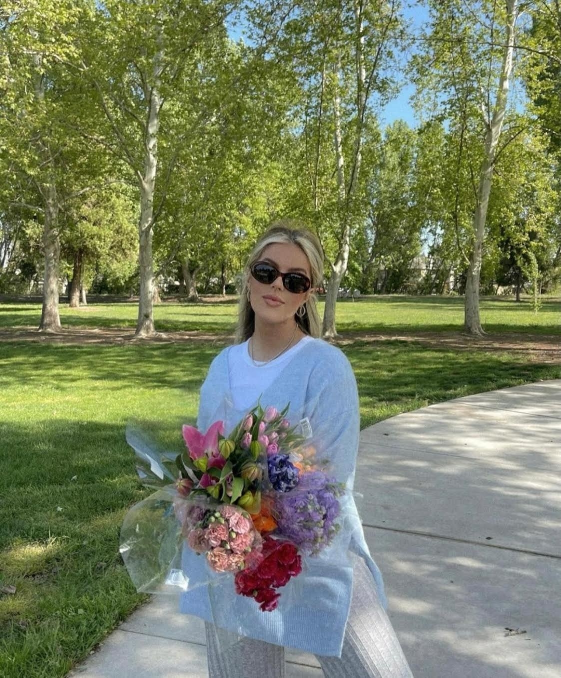 Woman in a park wears a sweater and holds a bouquet of flowers