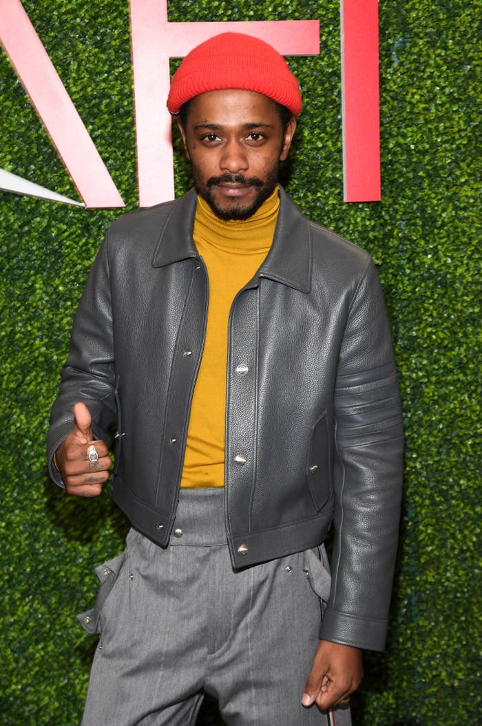 LaKeith wears a gray leather jacket, mustard high neck top, gray high waisted trousers, and a red fisherman&#x27;s hat