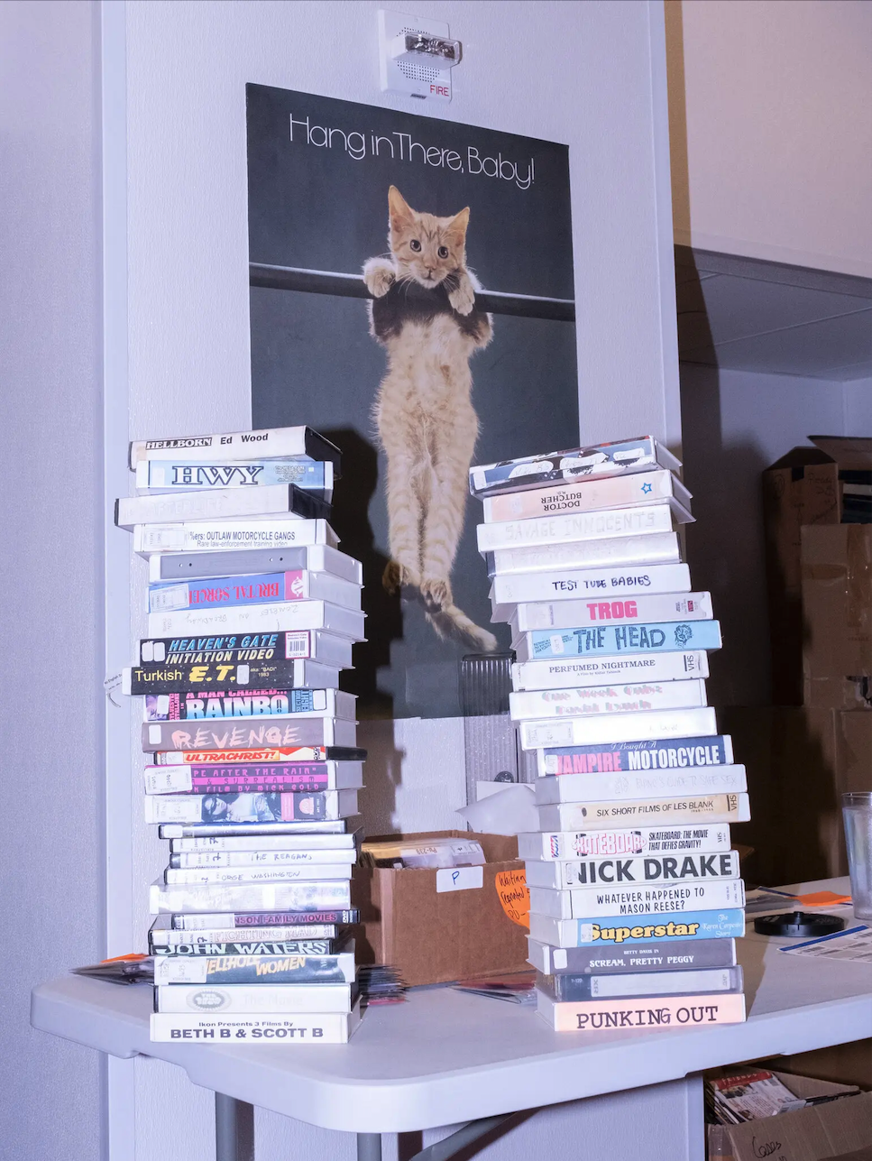 A &quot;Hang in There, Baby&quot; poster showing a kitten hanging on to a pole appears on a wall behind two stacks of VHS videotapes on a table