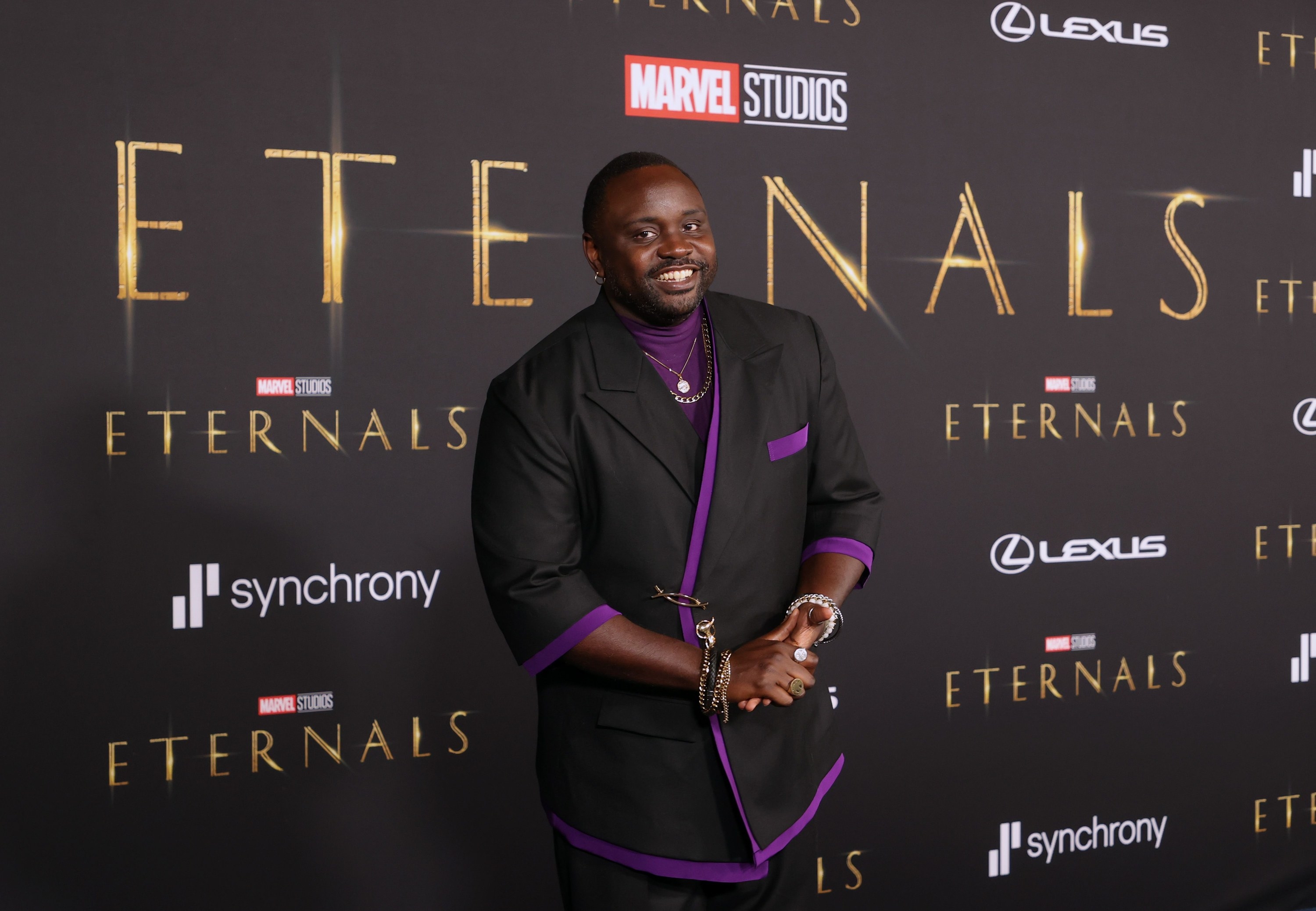 Brian smiles at the Eternals premiere, wearing an elbow-length black blazer with purple trim and a purple turtleneck