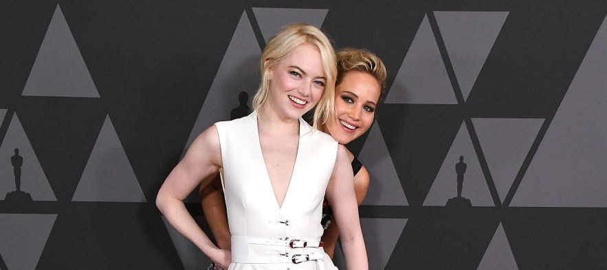 Jennifer smiling and standing behind a smiling Emma on the red carpet