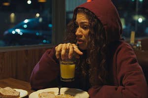 Rue from Euphoria sitting at a diner table with toast in front of her and a glass of orange juice in her hands