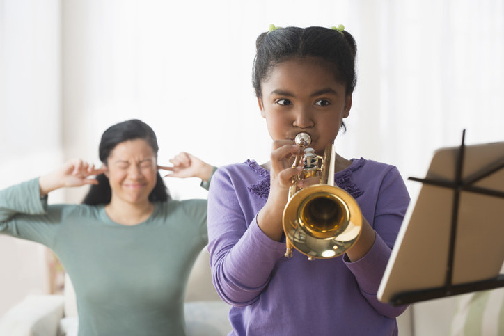 A girl playing the trumpet while her mother covers her ears in the background.
