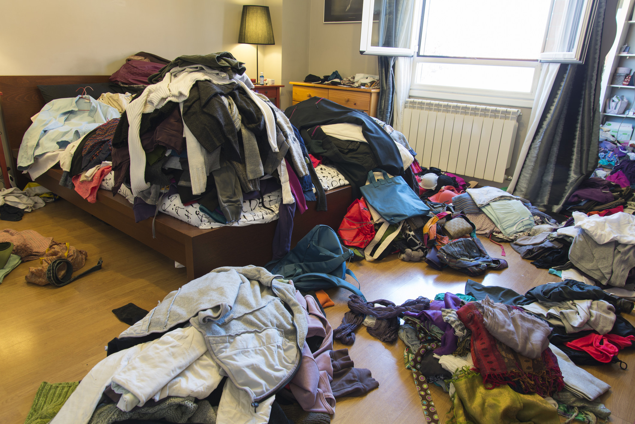 Wide angle view of large bedroom with piles of clothes on the floor and messy bed