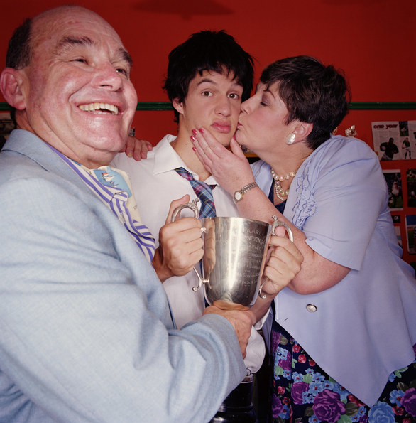 A mother kissing her son on the cheek while the father smiles and helps him hold up a trophy.