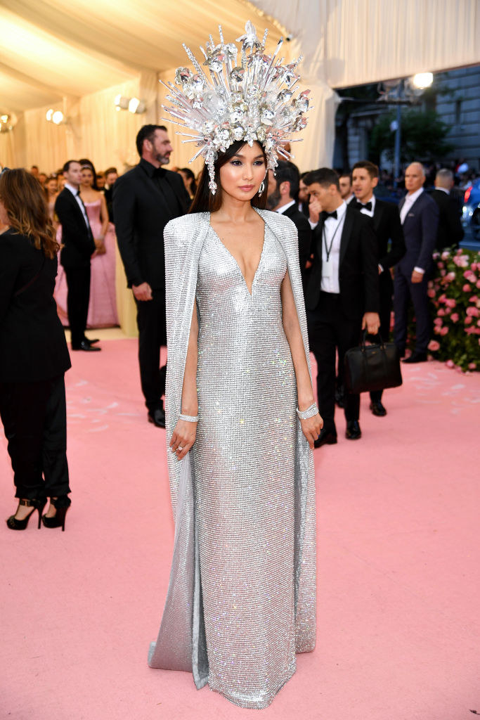Gemma Chan in a long sparkly evening gown, shawl, and crown