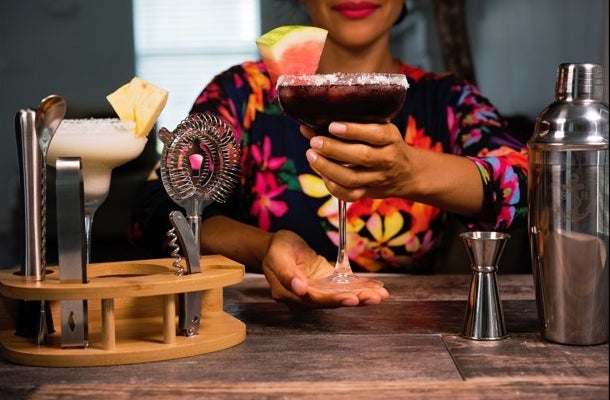 Someone holding a mixed drink next to the tools of the cocktail kit on the table