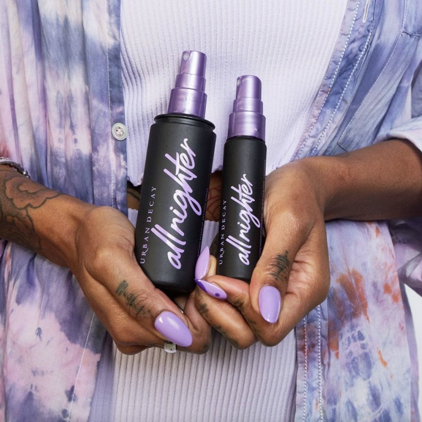 A person with a purple manicure, holding two bottle of makeup setting spray