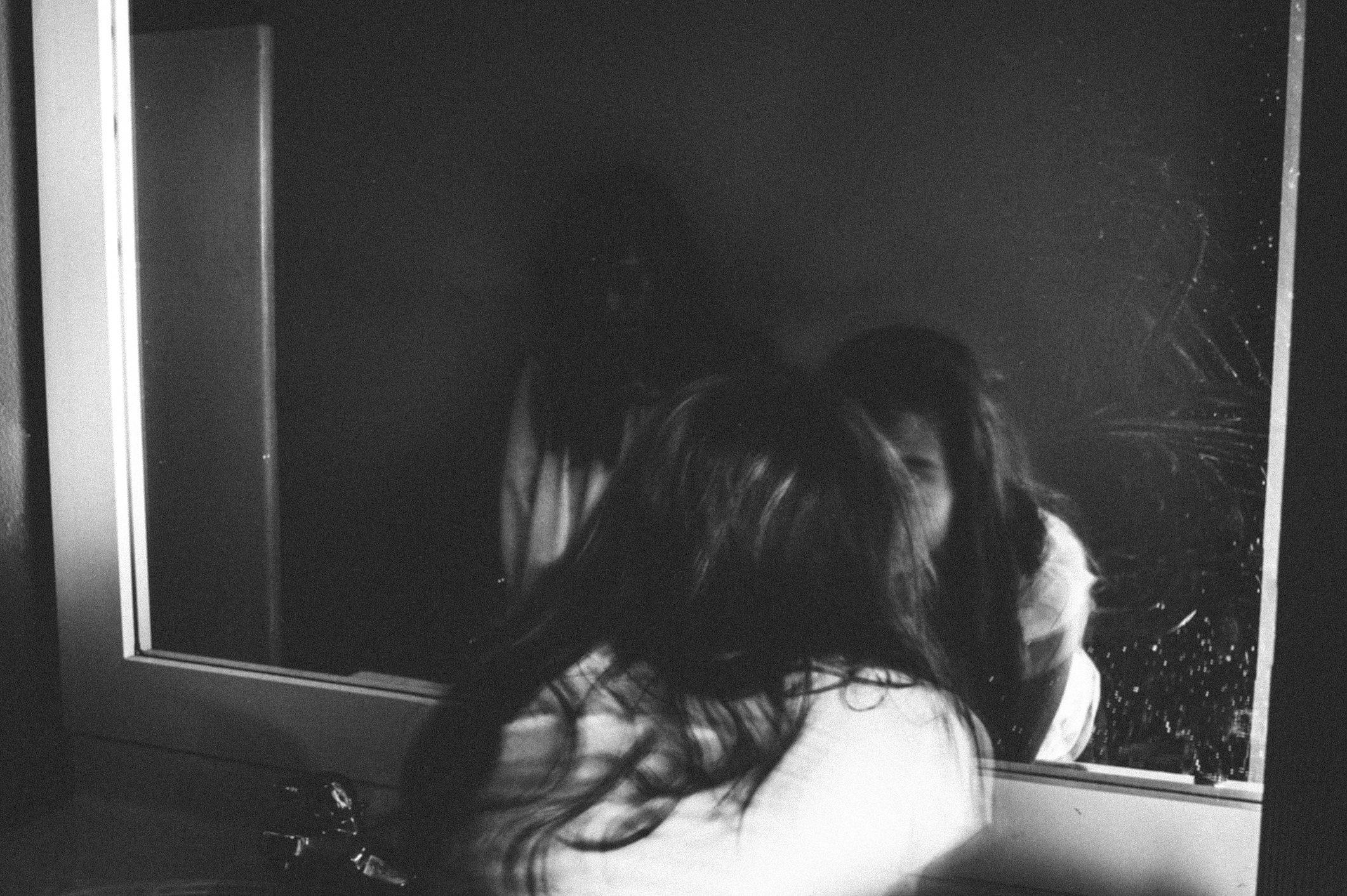 A girl leaning close to the mirror as a shadowy figure looks from behind her