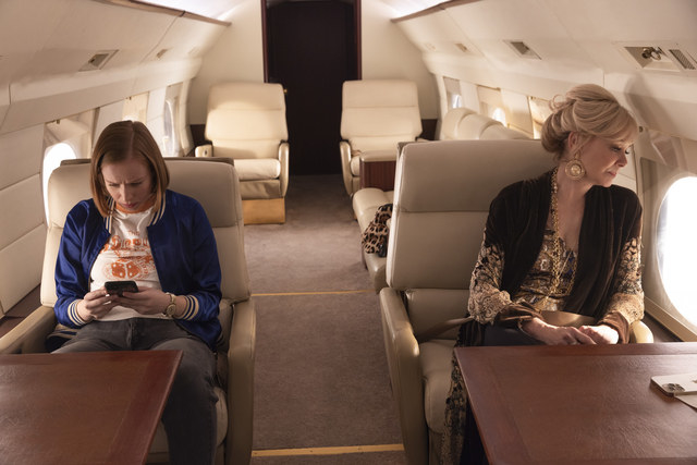 A young woman looks worryingly at her phone on a private plane while an older woman sitting across from her looks out the window