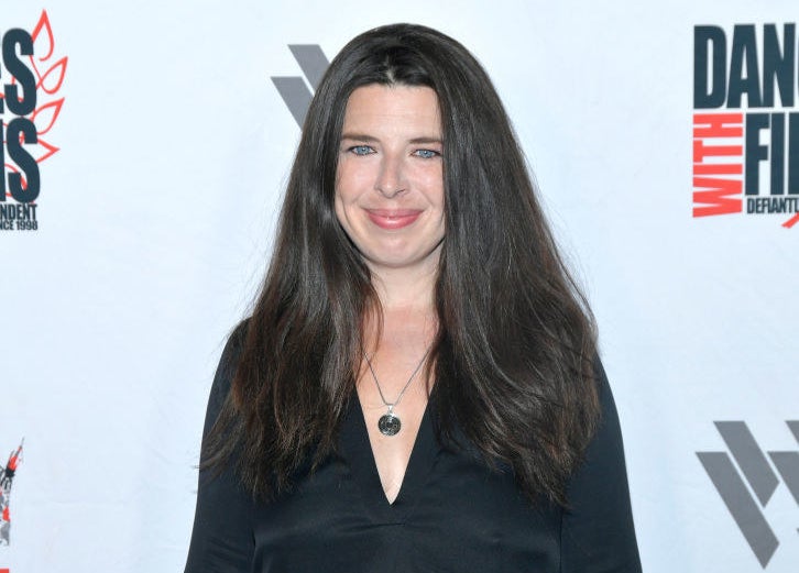Person on a red carpet wearing a deep V-neck black dress, smiling at the camera