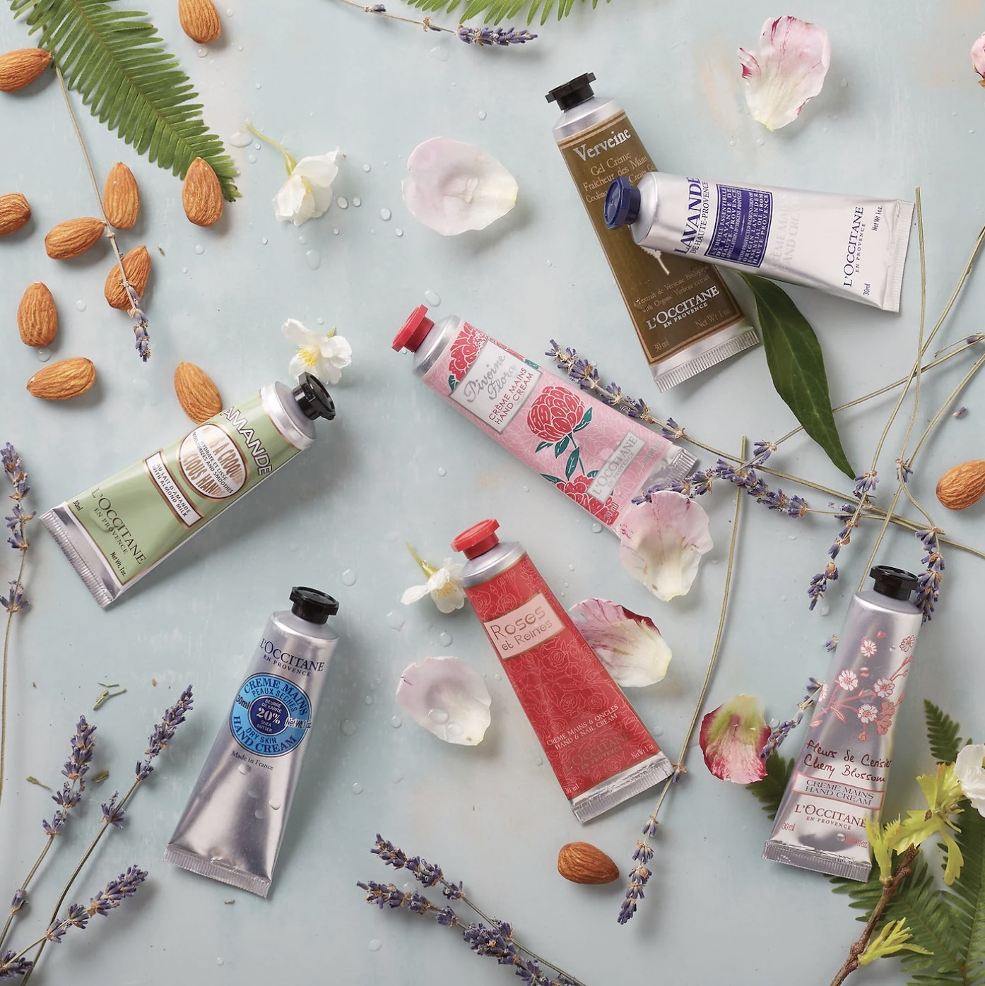 Tubes of the different scents of hand cream spread on counter with lilac stems, almonds and rose petals