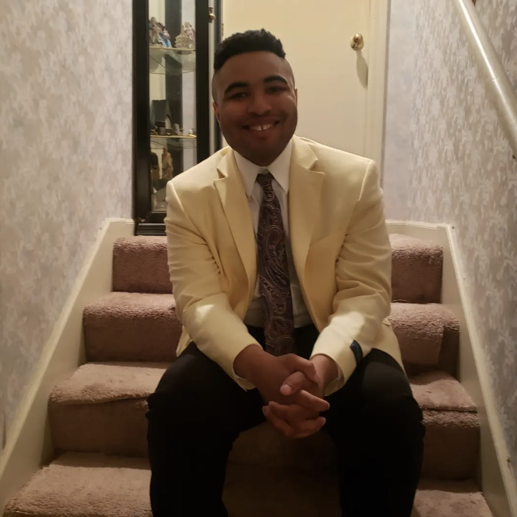 a man in a tie and blazer and sitting on carpeted stairs smiles for the camera 