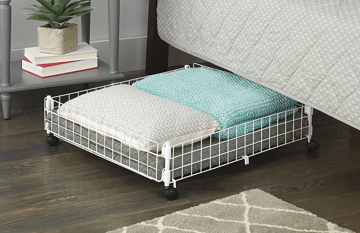 A wiry square basket on wheels peaking out from beneath a bed with pillows and blankets inside
