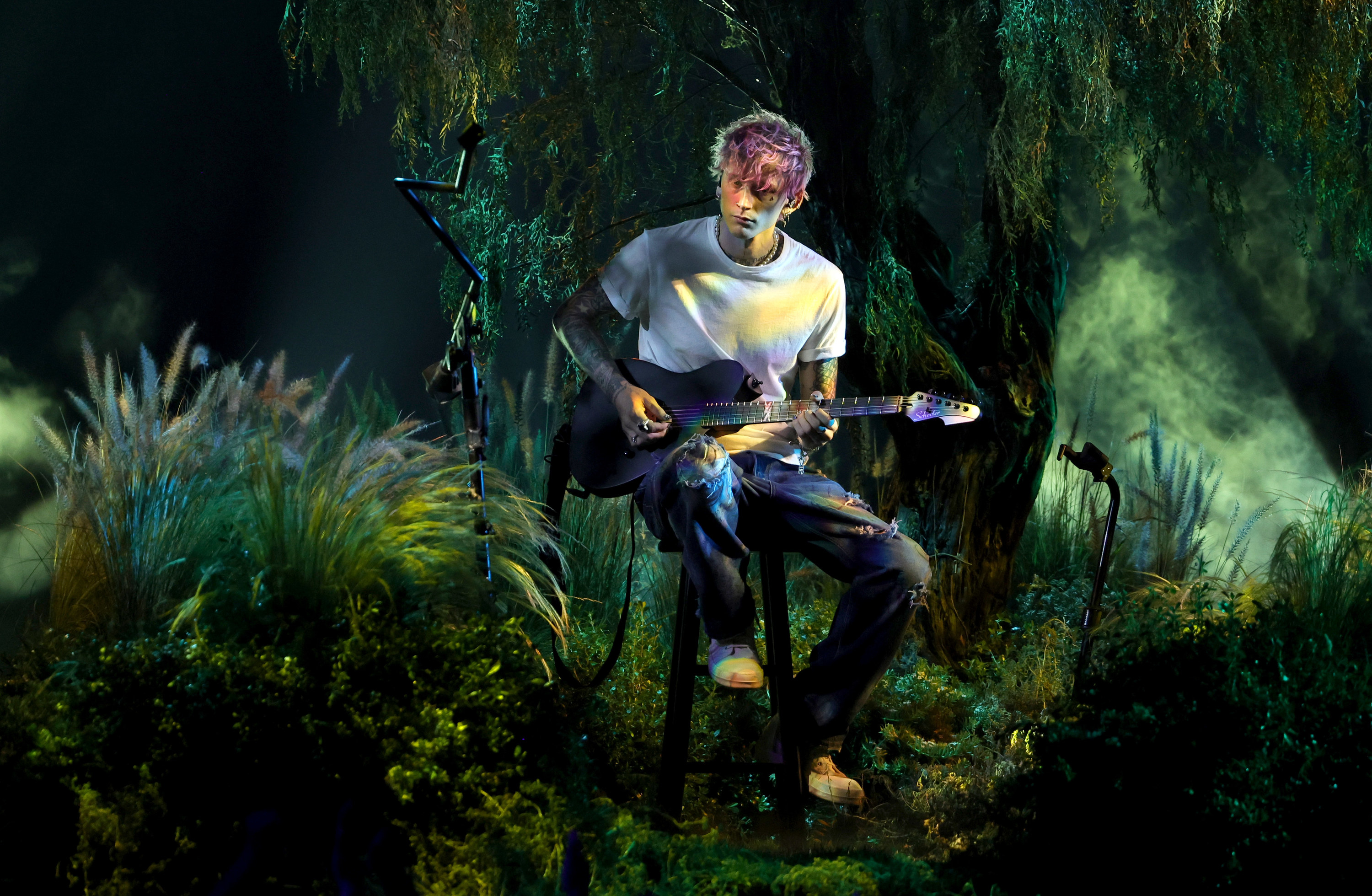 MGK sitting on a stool and performing against a backdrop of greenery