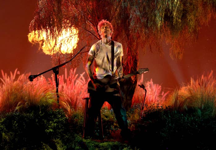 MGK performing with a guitar against a backdrop showing trees, grass, and a sun
