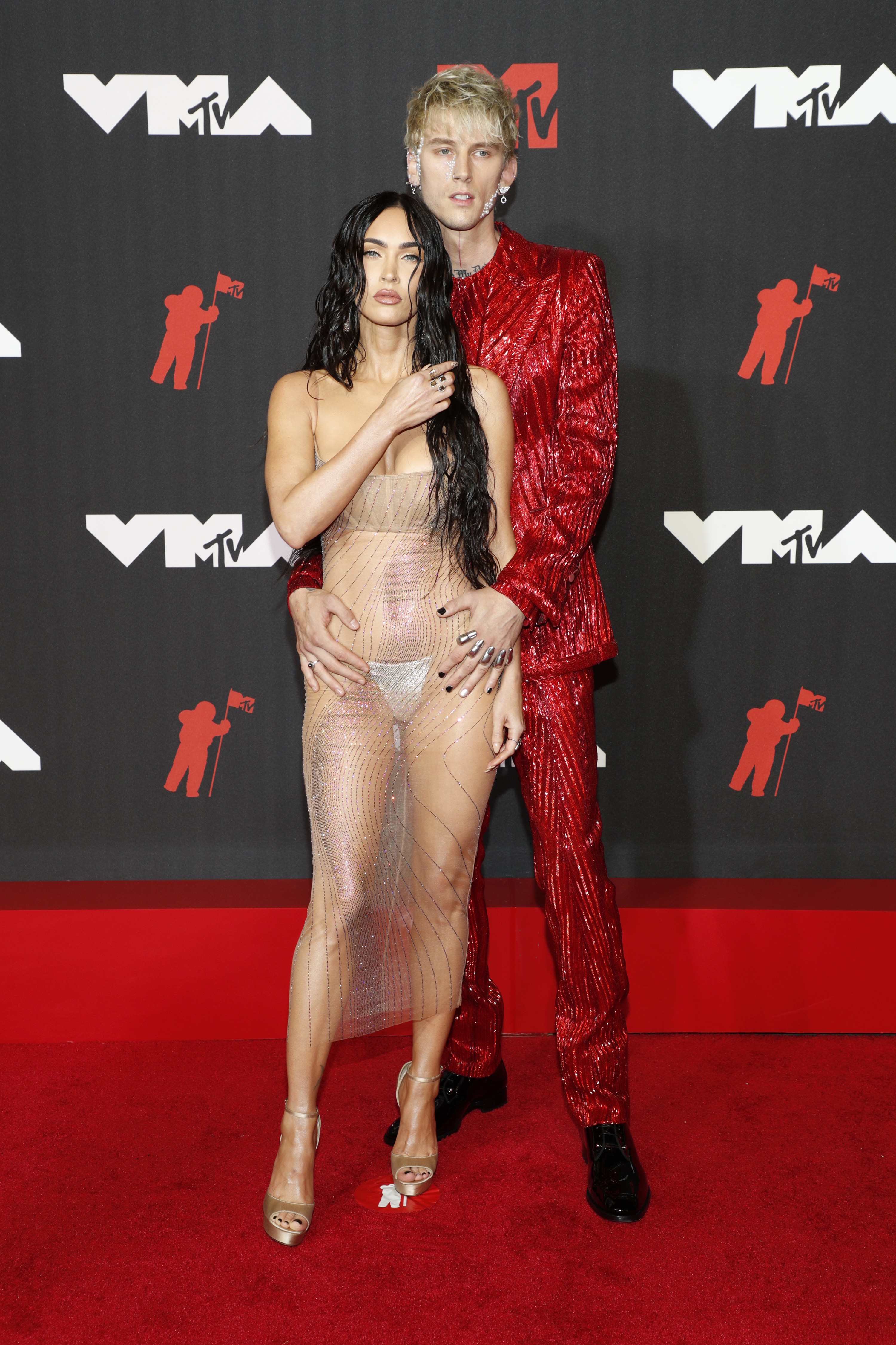 MGK standing behind Megan, who&#x27;s in a sheer gown, with his hands on her hips on the red carpet
