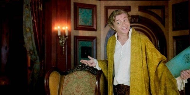 Stede Bonnet is in his captain's quarters, smiling and gesturing broadly with a book in his left hand