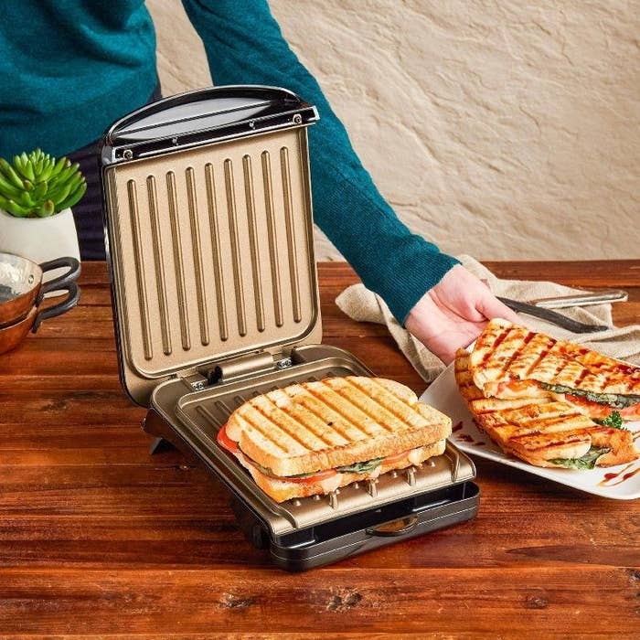 The panini press open with a grilled sandwich on the bottom plate while someone serves up a plate with another grilled sandwich on it