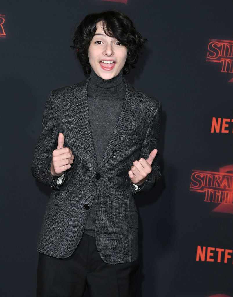 Finn throwing two thumbs up in a blazer and pant combo with long hair