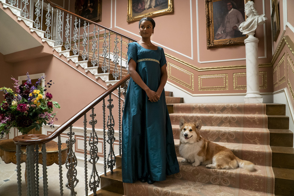 Simone as Kate standing on a sumptuous staircase with a dog on one of the steps
