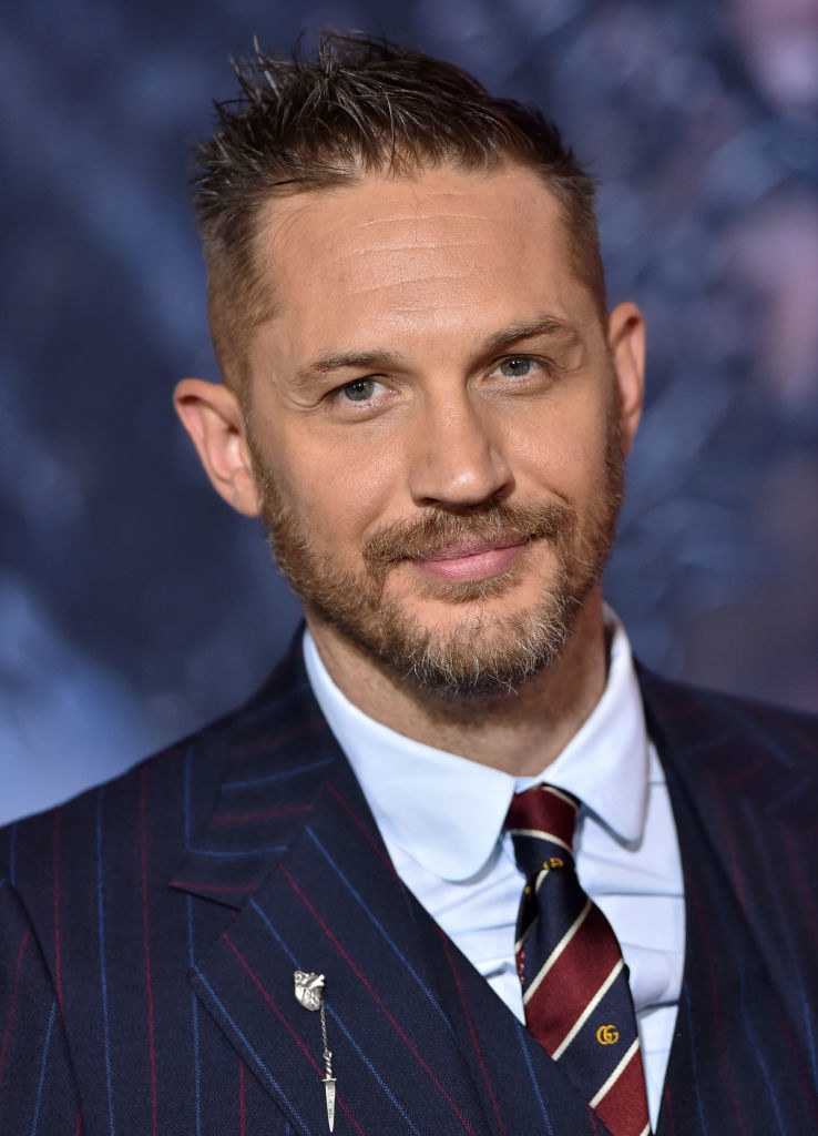 close up of Hardy with facial hair and spiked hair wearing a pinstripe suit