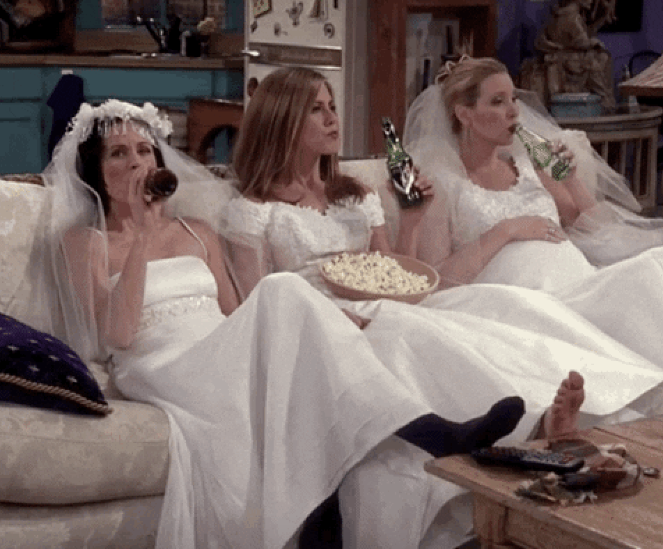 Rachel, Monica, and Phoebe from Friends sitting on a couch in bridal gowns, drinking beer, and eating popcorn