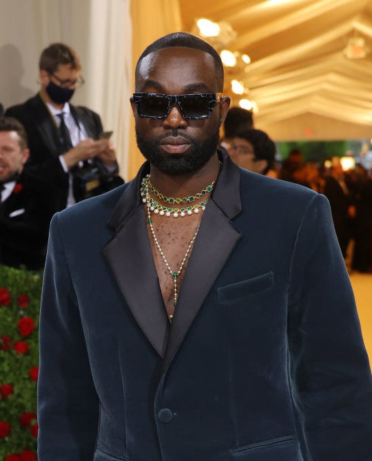 A closeup of Paapa Essiedu from the waist up that shows his emerald and pearl jewelry