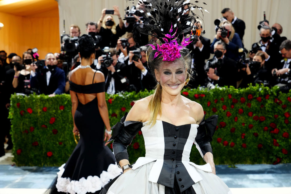 Sarah Jessica Parker smiles and wears a feathery hat and plaid gown