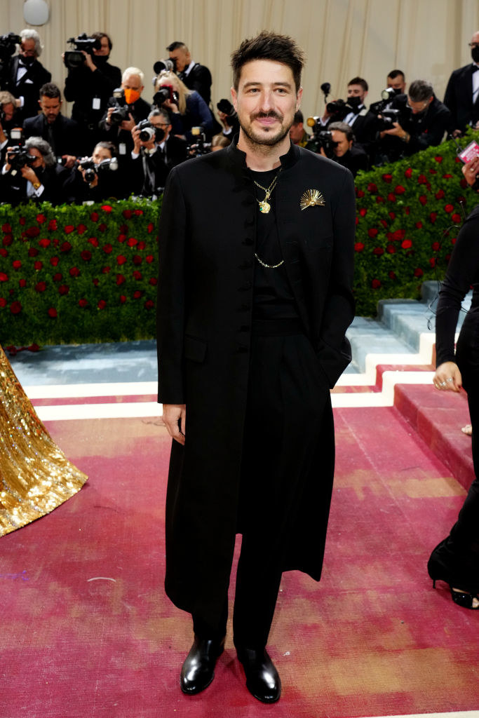 Marcus Mumford in a dark outfit with gold jewelry and a long coat