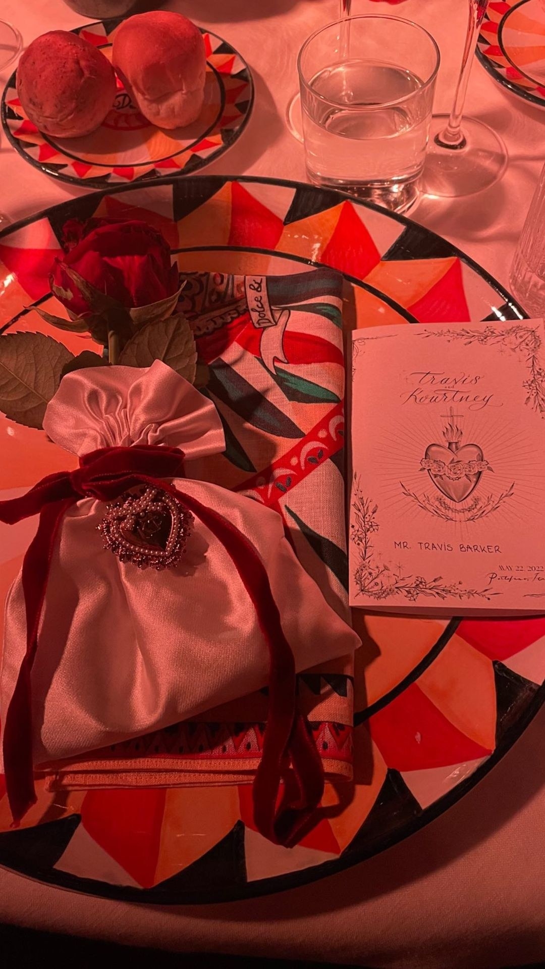 A party favor bag sits next to a place setting; the bag is wrapped with ribbon with a pearl-encrusted heart hanging off it