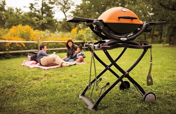 A gas grill with an orange top and a family in the background on a blanket