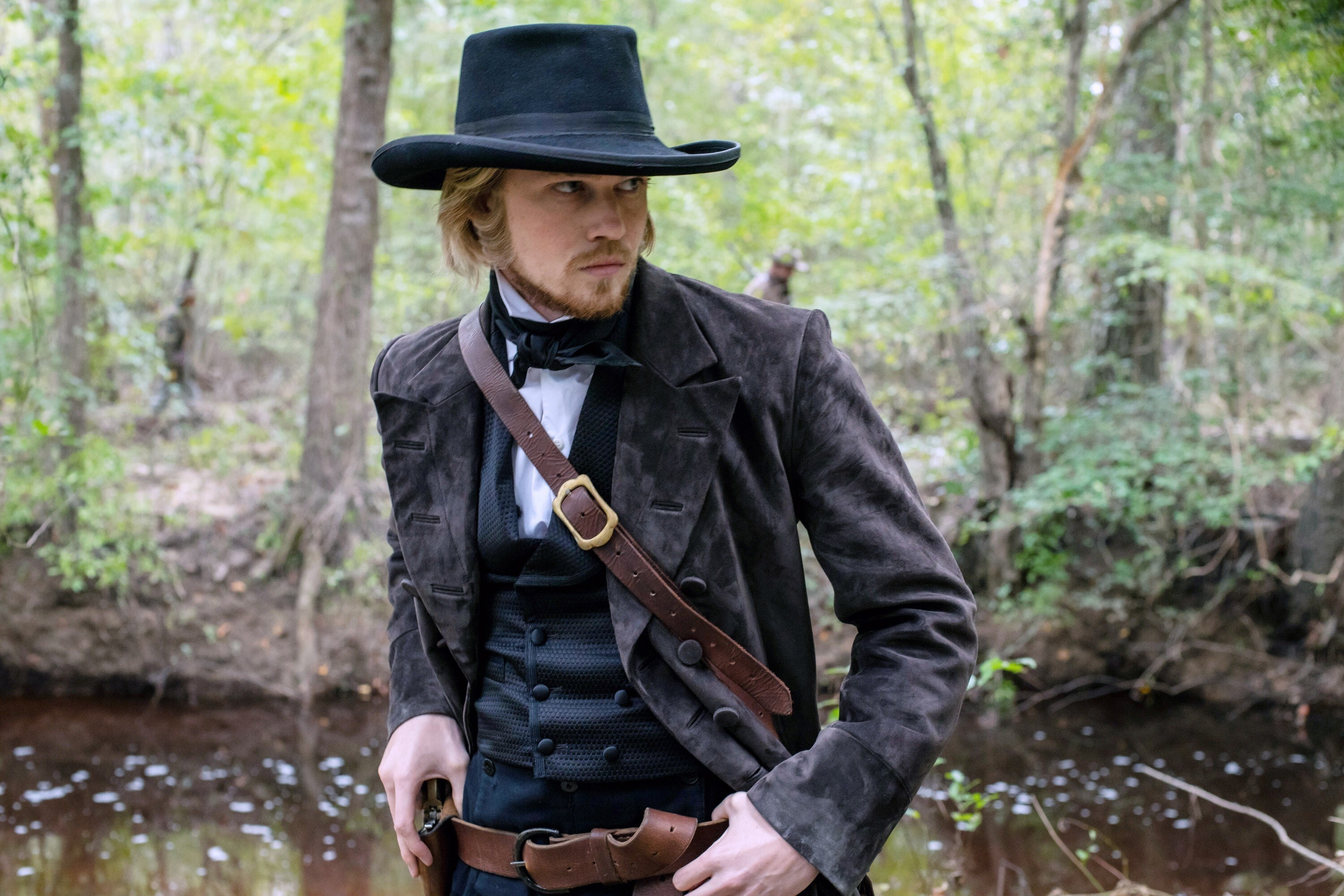 Joe Alwyn stands outside in front of a river; he wears a cowboy hat and has his hand on a gun holster