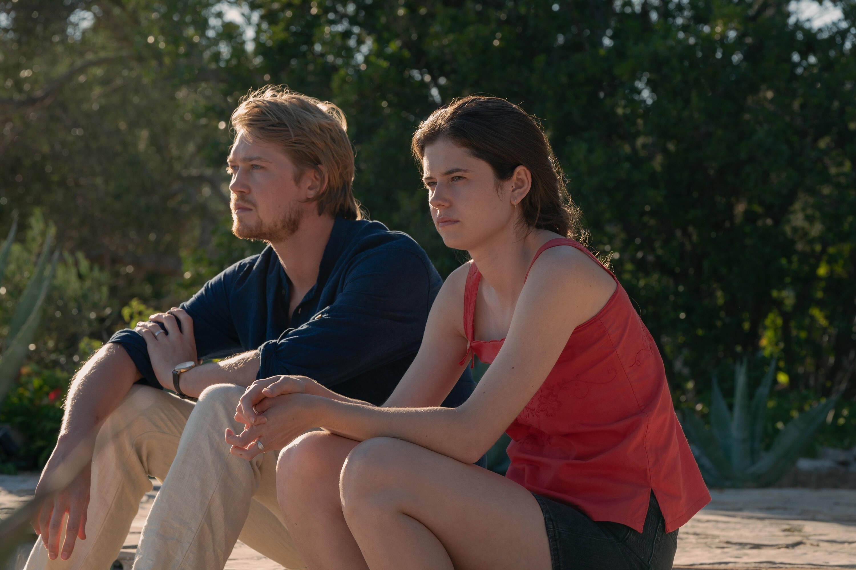 Joe Alwyn sits on the ground outside; a young woman sits next to him
