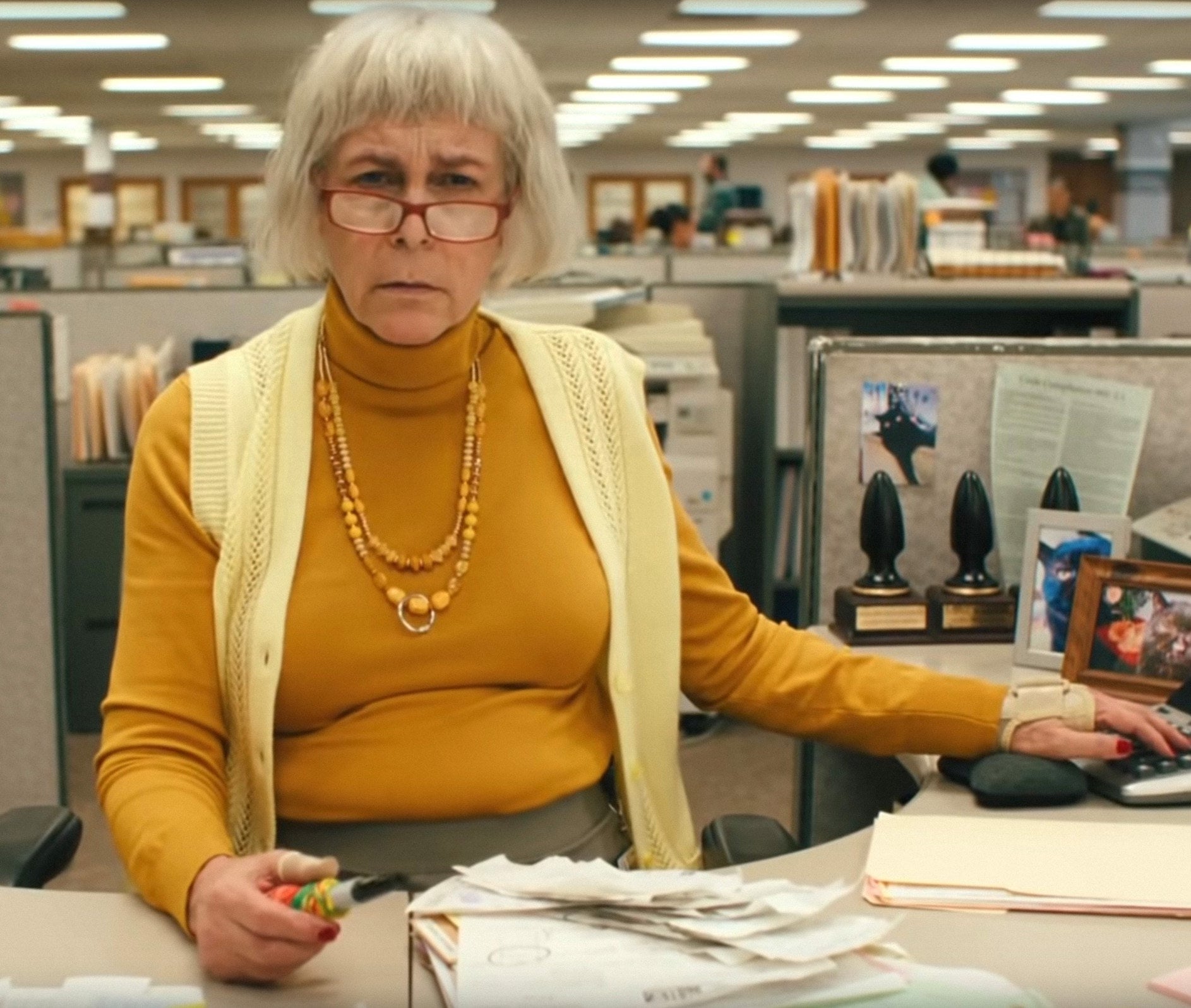 Jamie has a grey bob hair cut and wears a sweater vest over a turtleneck behind her desk