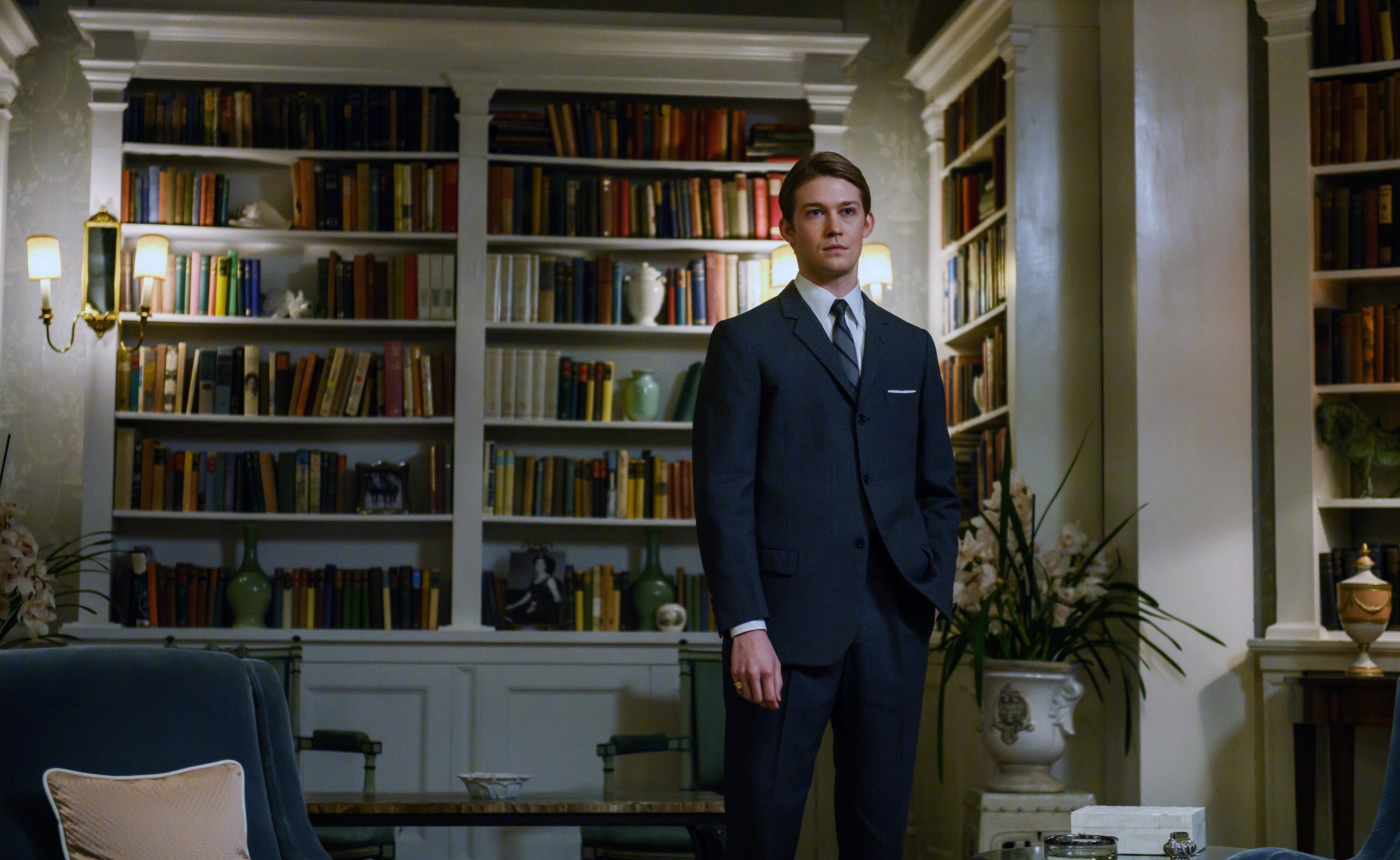 Joe Alwyn stands in front of a big book shelf wearing a suit