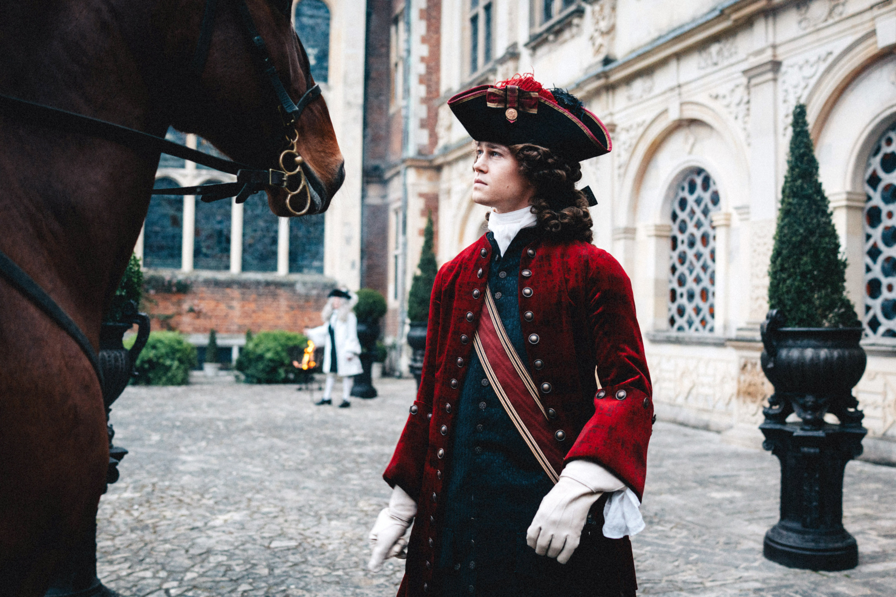 Joe Alwyn, dressed in 18th-century clothes, wig, and hat, stands outside in front of a brown horse