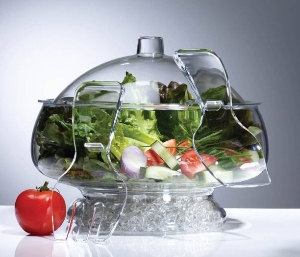 The glass bowl with ice in the bottom and a greens salad in the top portion covered by the dome lid with matching tongs hanging from side