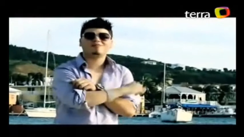Farruko wearing sunglasses and with his arms crossed against a backdrop of water, boats, and hills