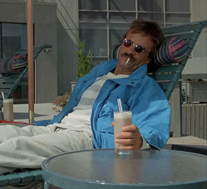 Terry Kiser in a blue jacket with sunglasses sitting on lounger with hand being lifted up