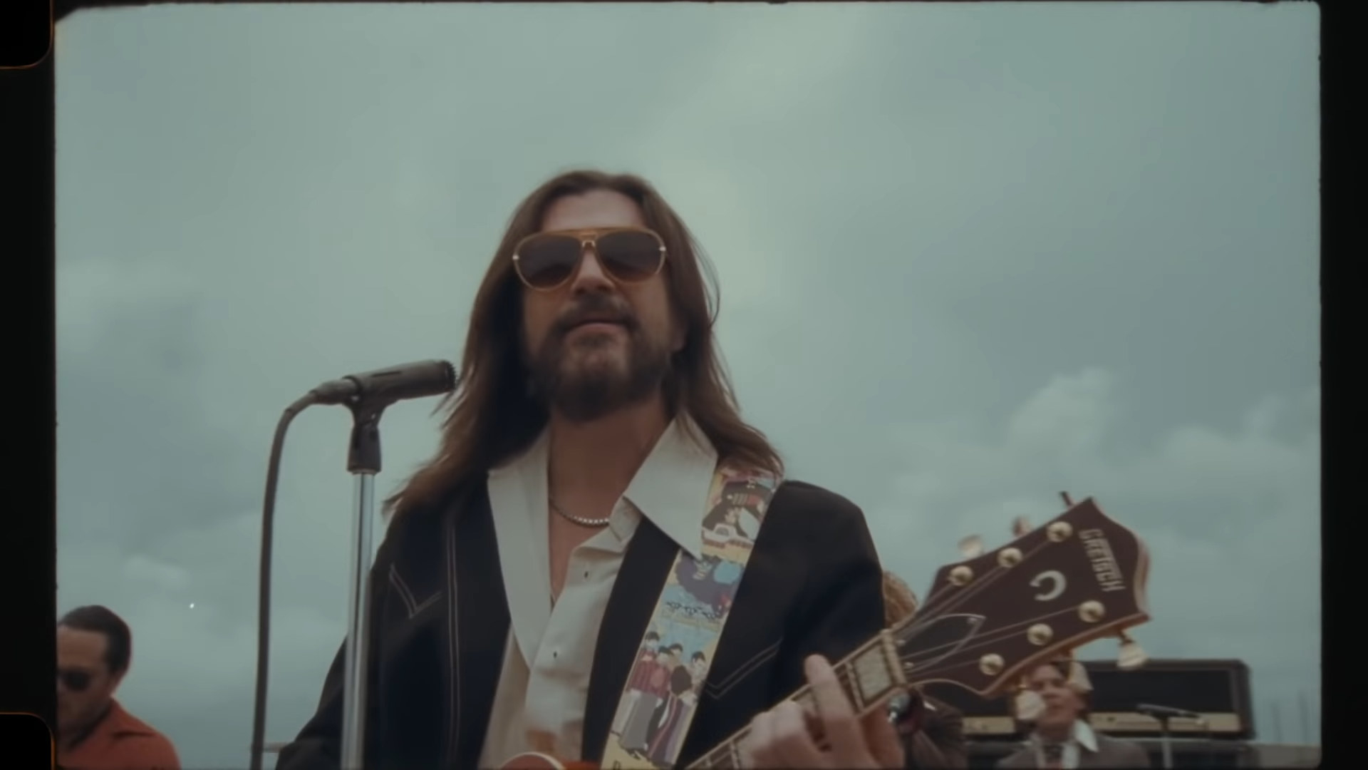 Juanes with long hair, wearing sunglasses, and playing a guitar at a microphone