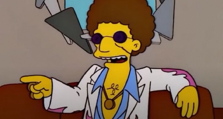 Disco Stu sits on a couch, he has a curly brown afro, a open shirt, a pink and white jacket and sunglasses