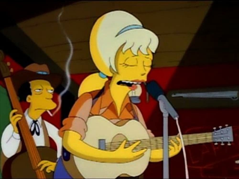 Lurleen Lumpkin, with a blonde beehive hairdo, stands on stage singing and playing guitar