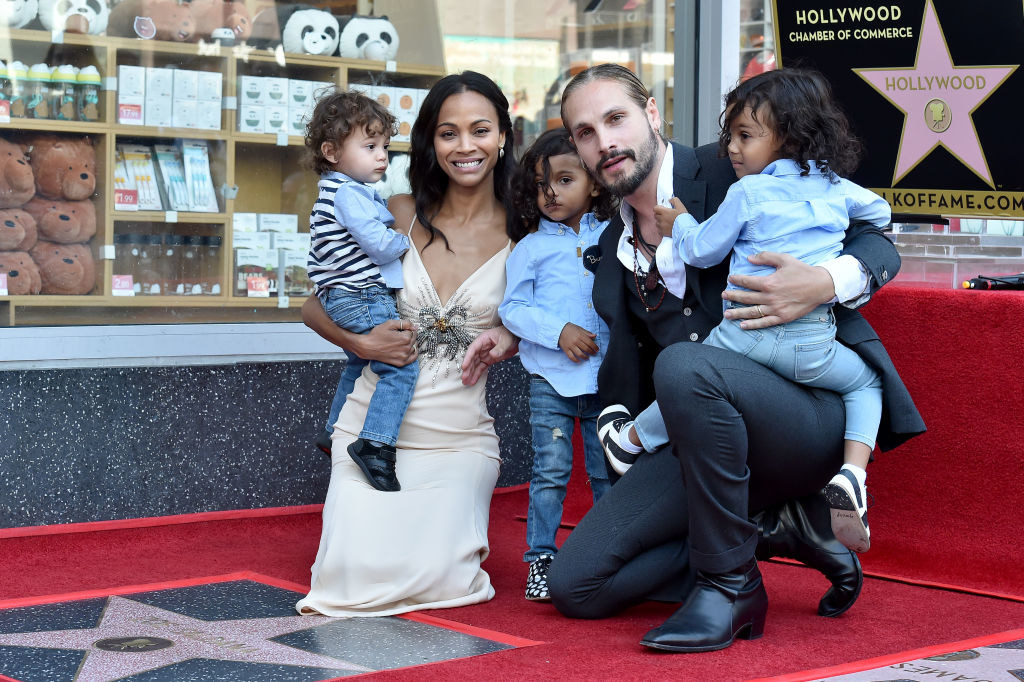 Zoe Saldana wears a light colored slip dress, Marco Perego wears a dark suit, and their three children wear light colored button up shirts with jeans