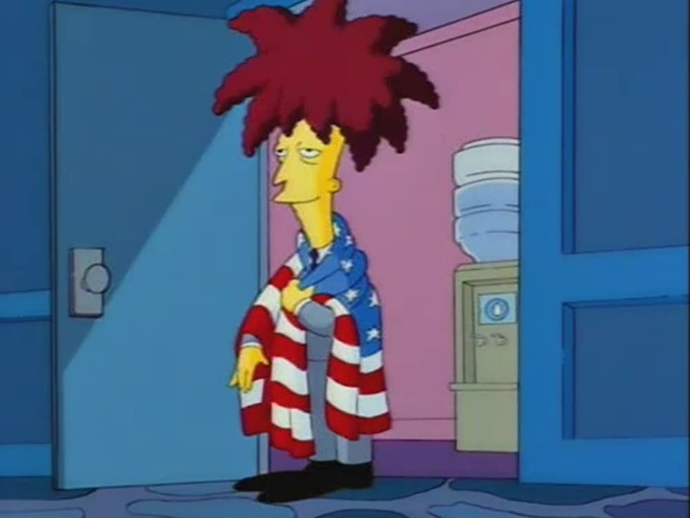 Sideshow Bob, with a giant red spiky afro, stands in a doorway wearing a suit and an American flag draped around him