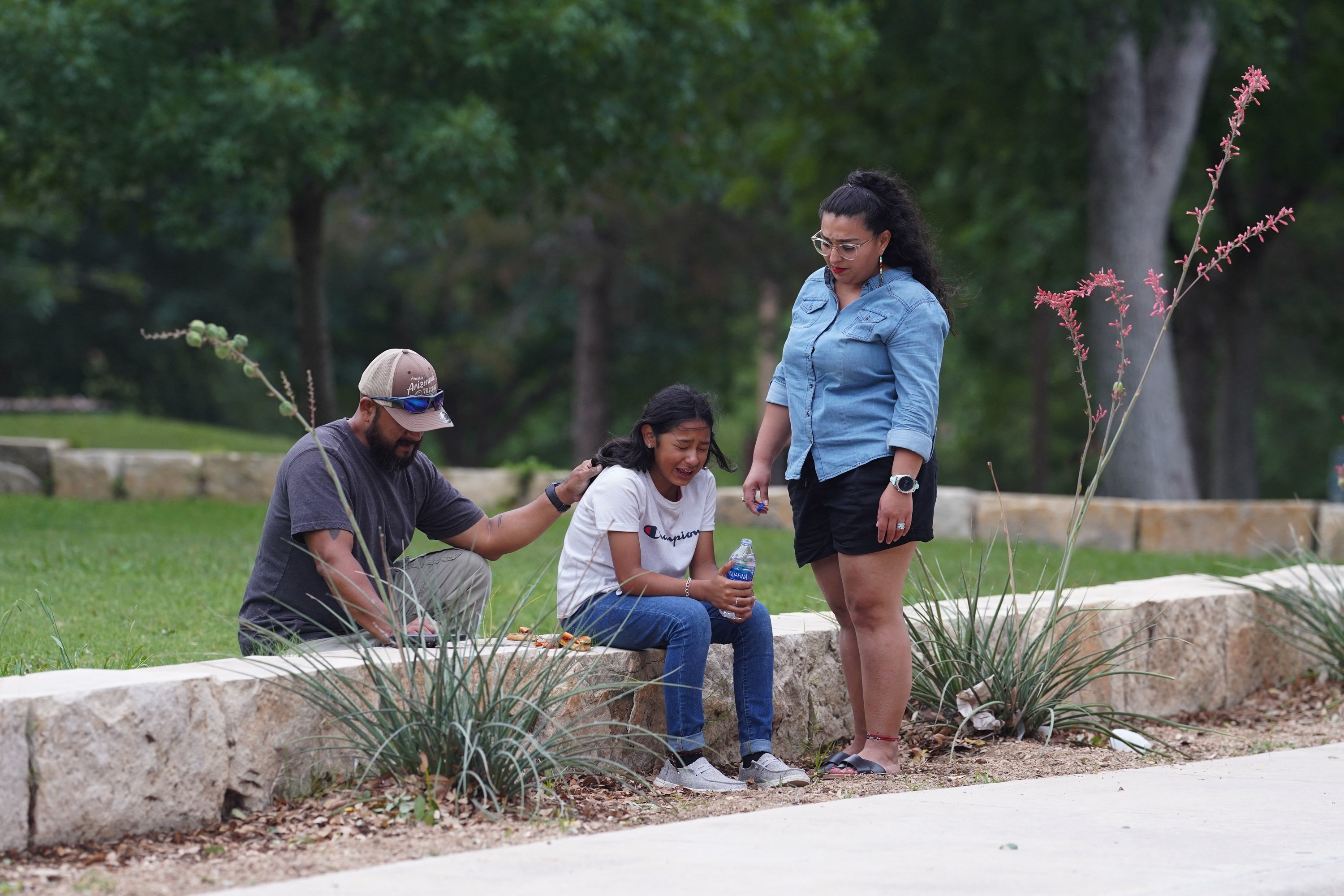 A young girl sits outside and cries as a man kneels behind her and places a hand on her back and a woman stands over here